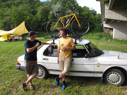 Right side view of a white car in a grass field, with 2 people leaning on it and a yellow Surly bike mounted on the roof